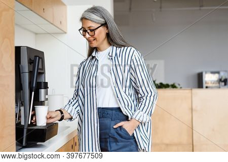 Happy white-haired mature woman smiling while using coffee maker indoors
