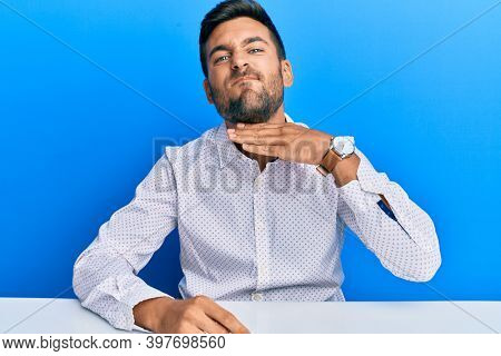 Handsome hispanic man wearing business clothes sitting on the table cutting throat with hand as knife, threaten aggression with furious violence 