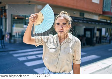 Young beautiful blonde caucasian woman smiling happy outdoors on a sunny day using handfan for hot weather
