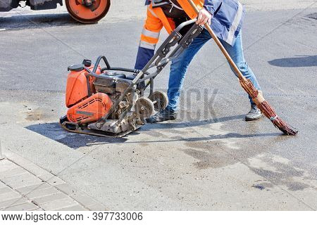 A Road Worker Adjusts The Operation Of A Petrol Vibratory Compactor Against The Background Of A Vibr