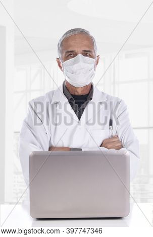 Docor wearing surgical mask standing behind a laptop computer in a modern clinic setting. 