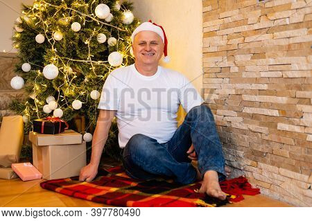 Grandfather Is Sitting Near Cristmas Tree In Santa Clauss Hat At Night At Home.