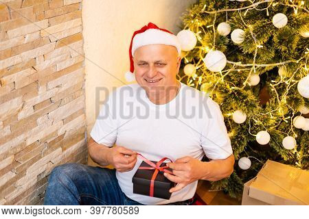 Grandfather Is Sitting Near Cristmas Tree In Santa Clauss Hat At Night At Home.