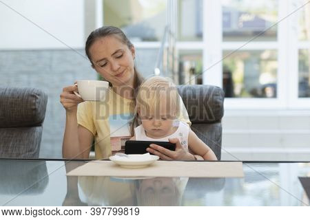 Young Mother In Cafe With Child In Her Arms Drinks Coffee And Looks At Phone. Modern Business Mom.