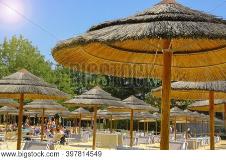 Straw Beach Umbrellas On The Beach Close-up Across Sunlight, Blu Sky, Green Plants And Vacationists,