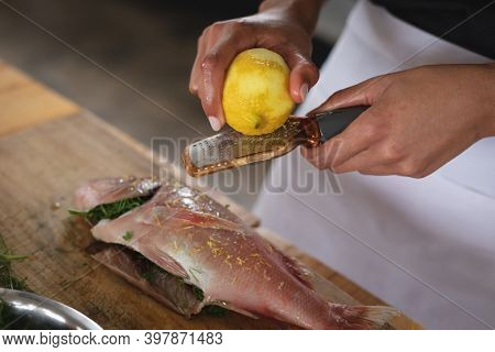 Mid section of chef grating lemon rind on roasted fish in restaurant kitchen. food preparation restaurant culinary gourmet