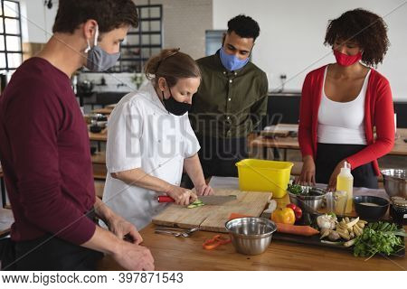 Female chef and divserse group wearing face masks in kitchen. trainee chefs standing around a table listening to female chef. hygiene in professional kitchen during coronavirus covid 19 pandemic.