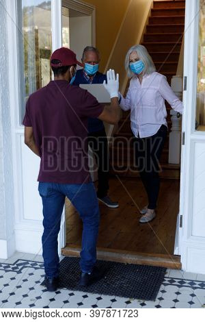Mid section of delivery man wearing face mask delivering package to senior caucasian couple wearing face mask at home. social distancing quarantine lockdown during coronavirus pandemic