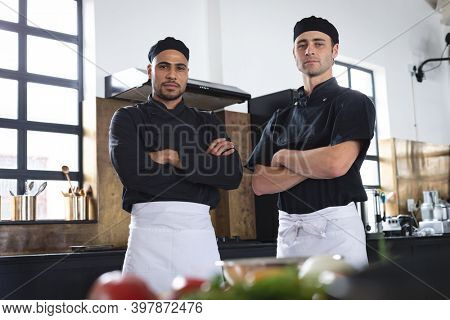 Two divserse male chefs in kitchen. portrait of two trainee chefs looking at camera with arms crossed.