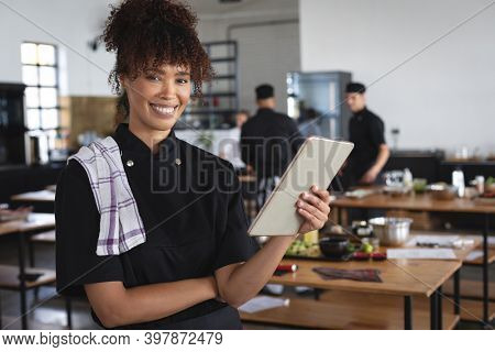 Mixed race female chef in kitchen. portrait of female trainee chef looking at camera and smiling, holding tablet, with diverse group of trainee chefs in the background.
