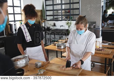 Female chef and divserse group wearing face masks in kitchen. trainee chefs standing around a table female looking at chef preparing food. hygiene in professional kitchen during coronavirus pandemic.