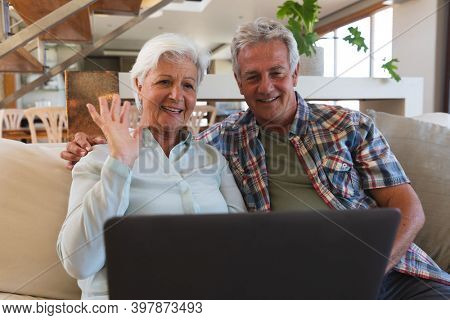 Senior caucasian couple on video call smilin and waving. sitting in livisng room at home using laptop. self isolation during coronavirus covid 19 pandemic