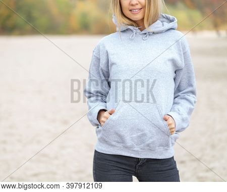 The front view of unrecognizable woman wearing light grey hoodie. She stands on the beach. Copy space on empty area on her blouse for design or inscription. Fashion mockup.