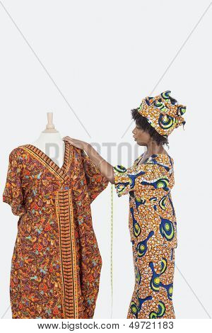 Female tailor in traditional outfit measuring dashiki on tailor's dummy over gray background