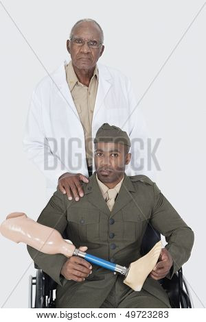 Portrait of senior doctor with US military officer holding artificial limb as he sits in wheelchair over gray background