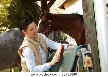 Young woman cleaning horse saddlery outdoors, brushing saddle-cloth.