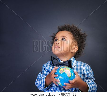 Portrait of cute little African boy holding in hands small globe on blackboard background, looking up, back to school concept