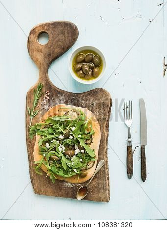 Rustic homemade pizza with eggpant, cheese, olives and arugula on rustic wooden serving board over g
