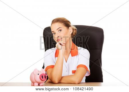 Young smile female doctor or nurse sitting behind the desk and holding a piggybank.