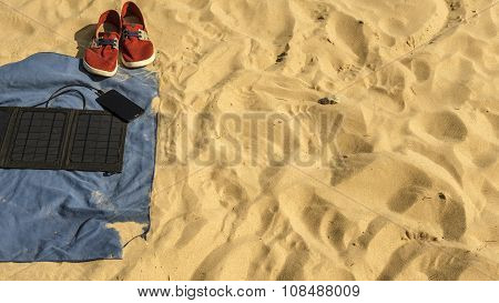 In The Sand On The Beach Are: Towel, Sneakers, A Phone That Is Charged From A Solar Panel Battery.