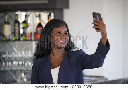 young attractive and happy black afro american woman in casual elegant clothes taking selfie portrait photo with mobile phone camera smiling cheerful and positive sitting at restaurant bar
