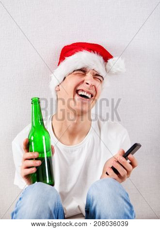 Cheerful Young Man in Santa Hat with Bottle of a Beer and Cellphone by the Wall