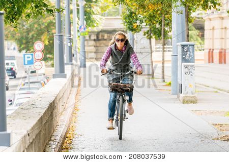 Budapest Hungary - September 27 2017: Selective focus front view of a caucasian woman riding a bicycle at an walk and cycle path in Budapest Hungary to avoid traffic. Autumn trees and traffic in the background.