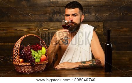 Viticulture and autumn harvest concept. Man with beard holds glass of wine on vintage wooden background. God Bacchus with interested face wearing white cloth sits by wine bottle and basket with fruit