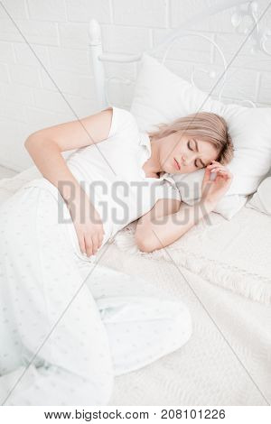 Blonde, young girl in white clothes lies on a bed with a pain in the stomach on a white background