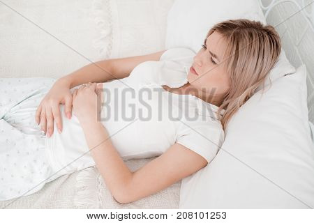 Blonde, young girl in white clothes lies on a bed with a pain in the stomach on a white background