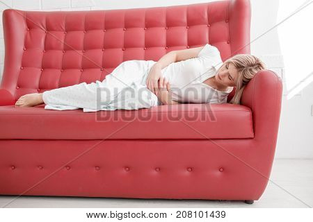 Blonde, young girl in white clothes lies on a red couch with pain in the stomach on a white background