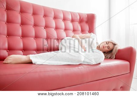 Blonde, young girl in white clothes lies on a red couch with pain in the stomach on a white background