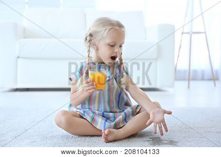 Cute little girl with glass of juice sitting on carpet near wet spot
