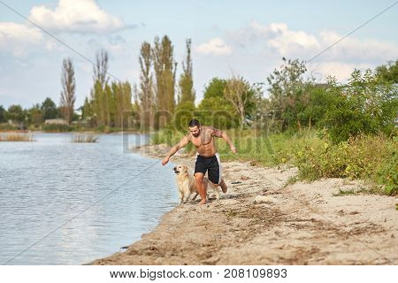 A young man with muscular body runs on the beach with his friend dog. An owner and dog walking on a nature background.