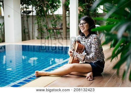 Young Woman Hold Guitar Classic In Her Hand And Sitting Beside Pool Around Her Has Peaceful Green Na
