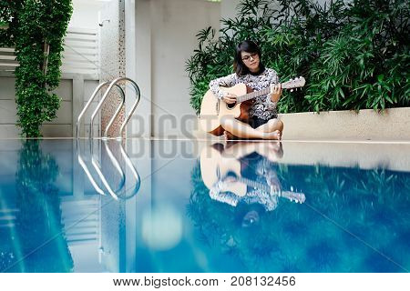 Young Woman Hold Guitar Classic In Her Hand And Sitting Beside Pool Around Her Has Peaceful Green Na