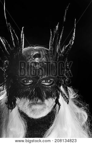 Male bearded face with long white hair and ominous sight in vintage decorative masque with dark brown feathers for fancy ball on black background studio