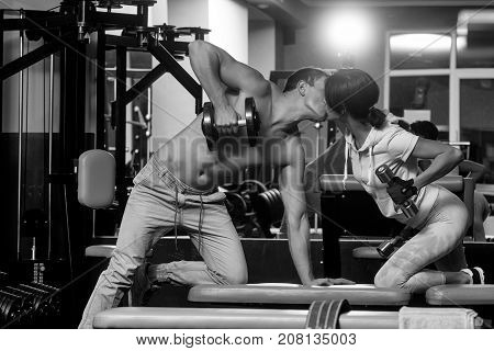 kissing couple of pretty woman or cute girl and handsome guy or macho man with muscular torso and sexy body training in sport gym with dumbbells at trainer of weight lifting
