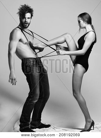 Pretty girl or sexy woman in black bodysuit and red high heel shoes pulling suspenders of bearded man with beard and muscular torso in jeans on grey background