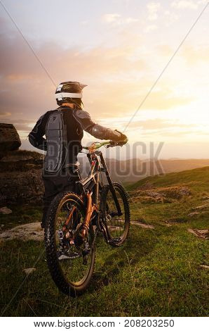 Ryder in full protective equipment on the mtb of two pendant bicycles rides along the green grass towards the sunset in the rays of the sunset sun against the backdrop of a landscape of rocky terrain under the setting sun and clouds.