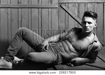 Handsome fashionable sexy sensual muscular young macho man with bare torso and stylish hair in jeans shirt indoor lying on wooden background horizontal picture