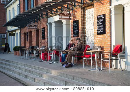 Luneburg Germany- January 28 2017: Street cafe with old couple drinking coffee at table outdoors on cement stairs on red brick wall background. Leisure lifestyle enjoy life concept