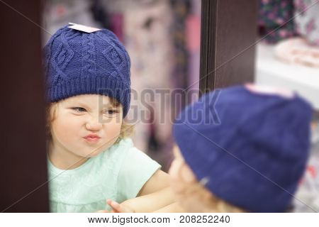 Cute Little Girl Choosing Clothes In The Shop, Fitting Knitted Hat.