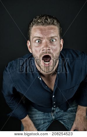 Macho with open mouth on bearded face leaning forward on dark background vintage filter. Man wearing fashionable blue shirt and jeans. Surprise and sensation concept.