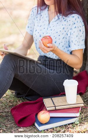 Close-up of a female student sitting next to textbooks, cup of hot coffee and apple on a blurred natural background. Modern student habits concept.
