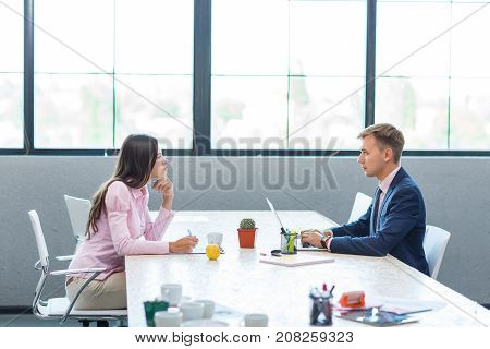 Young male colleagues and a woman sitting opposite each other in the office. Against the background of large windows. They look into each other's eyes.