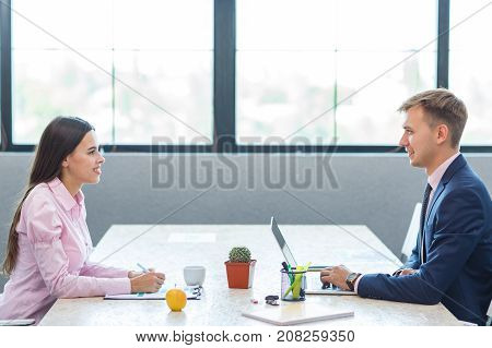 Young male colleagues and a woman sitting opposite each other in the office. Against the background of large windows. Smile and look into each other's eyes.