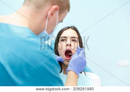 A dermatologist, a blonde man in purple gloves, examines the teeth of a young pretty black-haired girl at his reception