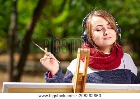 Beautiful girl draws on an easel and listens to music with closed eyes