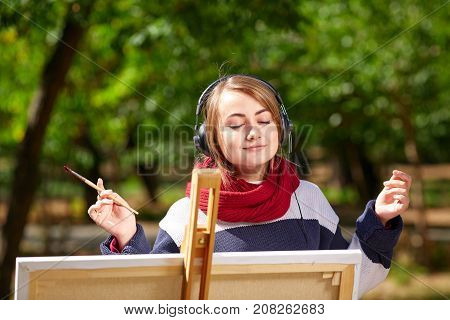 Beautiful girl draws on an easel and listens to music with closed eyes